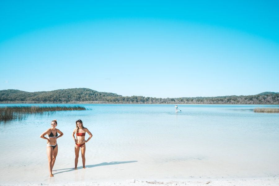 two girls standing next to a blue lake on K'gari