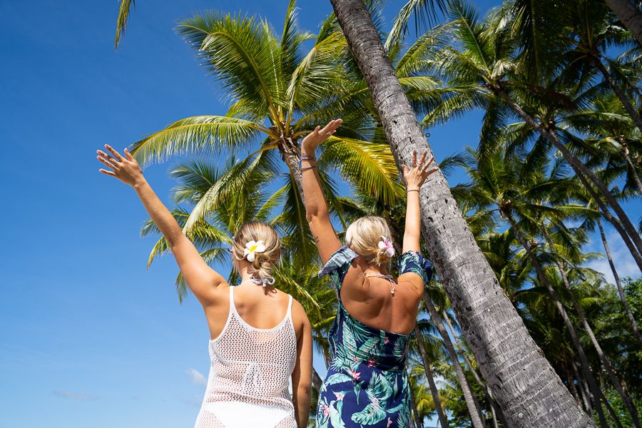 Two women with flowers in their hair and arms raised, palm trees in the distance