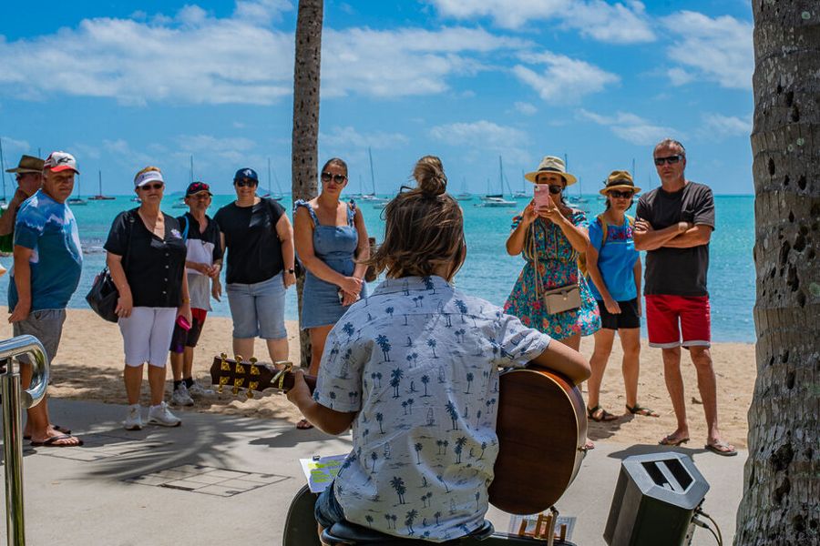 A man with his back to the camera playing a guitar facing a crowd in front of the beach 