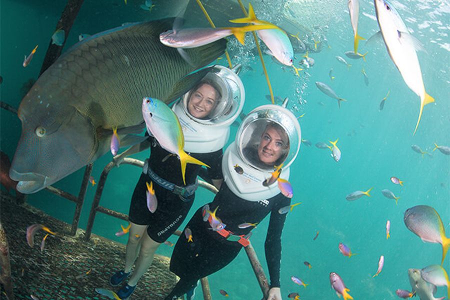 Two girls underwater wearing diving helmets surrounded by fish