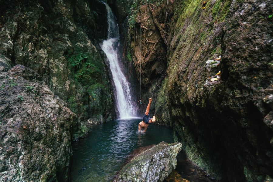 Man diving into Fairy Falls swimming hole from rock. Tall waterfall cascades in background, rope swing hangs from  moss rocks. 