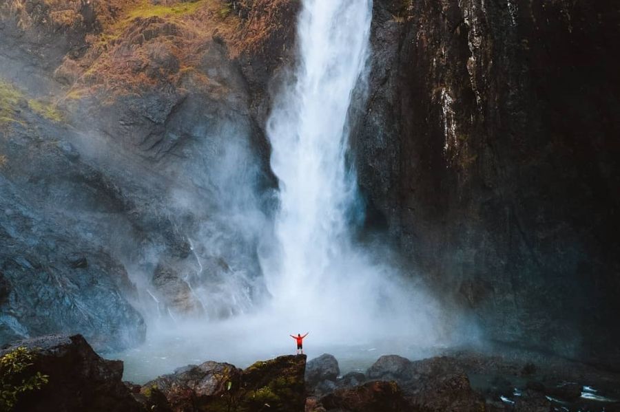 Clouds of mist at base of Wallaman Falls in background, silhouette of hiker raising hands toward waterfall in foreground