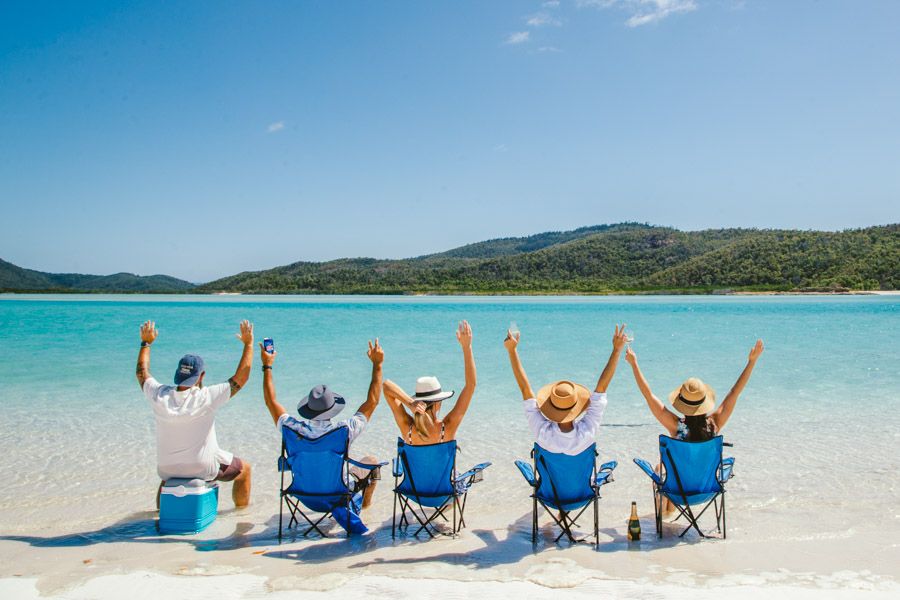 Five people sitting on chairs at Whitehaven Beach with their arms in the air