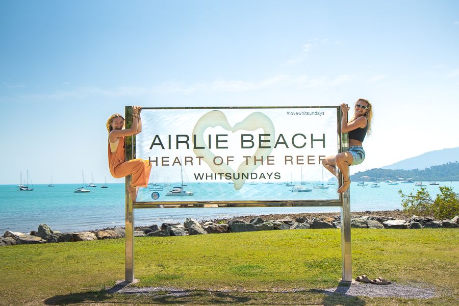 Airlie Beach two women hanging on to either side of a sign that says 'airlie beach'