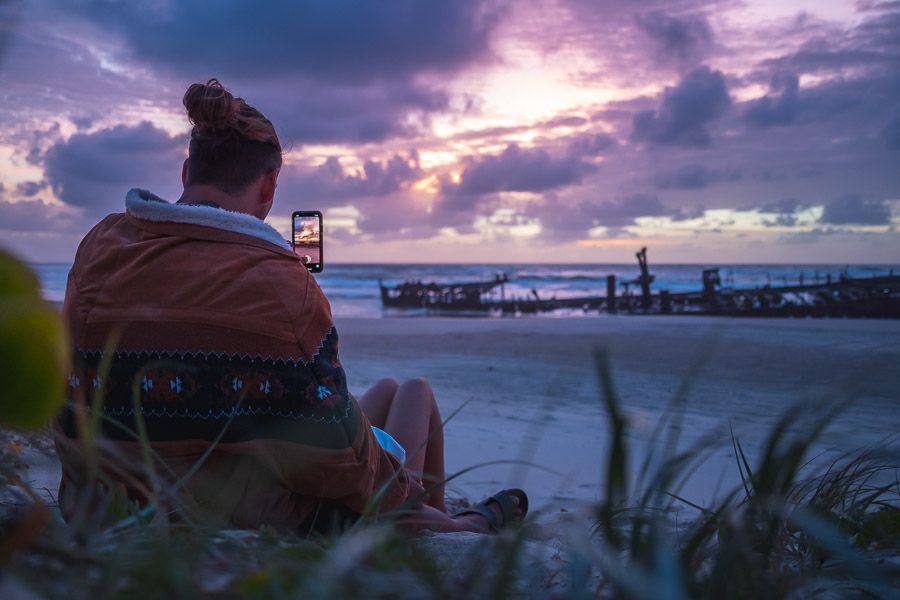 Boy sitting on dunes taking picture of Maheno Shipwreck at sunset