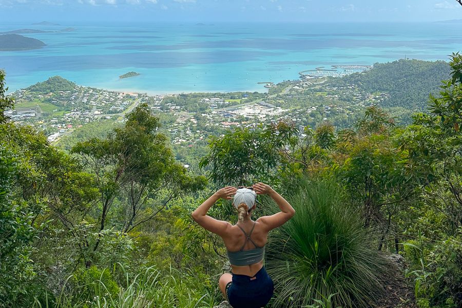 A girl sitting on the top of a mountain holding on to her hat looking at the view of a marina