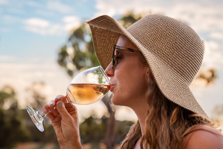 A woman sipping a glass of wine with a wide-brimmed hat and sunglasses on
