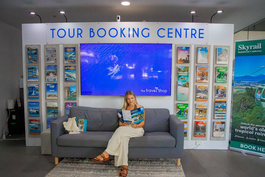 A woman holding a travel brochure sitting on a sofa under a sign that says tour booking centre