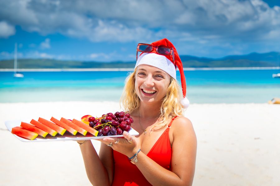 a woman wearing a santa hat holding a platter of food on the beach